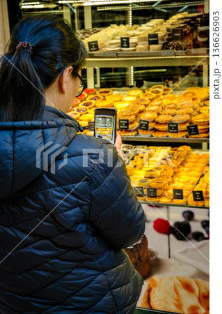 Anonymous woman in black puff jacket taking photo of traditional Portuguese pastries including famous pastel de nata in bakery window with smartphone. Food photography for social media blog. Tourist Anonymous woman in black puff jacket taking photo of traditional Portuguese pastries including famous pastel de nata in bakery window with smartphone. Food photography for social media blog. Tourist 136626903