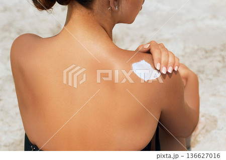 Attractive woman with healthy skin applying sunscreen to shoulder wearing white sun hat 136627016
