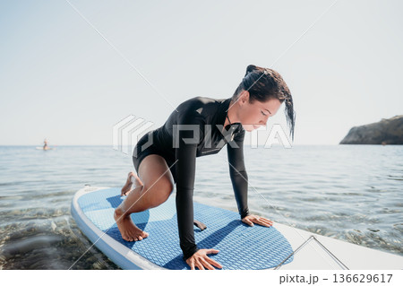 Yoga Paddleboard Water Workout Woman - A woman performs yoga poses while standing on a paddleboard in the water. She is wearing a wetsuit and appears to be enjoying the workout. The image is a perfect Yoga Paddleboard Water Workout Woman - A woman performs yoga poses while standing on a paddleboard in the water. She is wearing a wetsuit and appears to be enjoying the workout. The image is a perfect 136629617