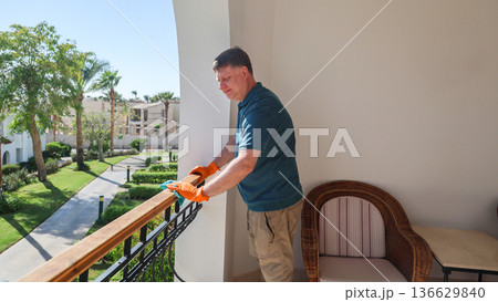 A middle-aged man wipes dust from the railing on a balcony 136629840