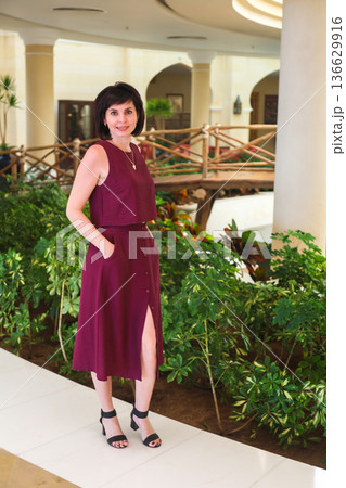Portrait of a brunette in a burgundy dress standing in a lobby near green plants Portrait of a brunette in a burgundy dress standing in a lobby near green plants 136629916