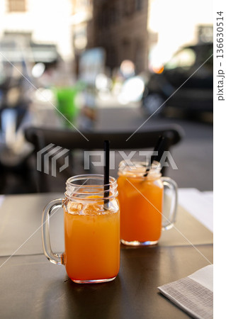 Two mason jars filled with fresh orange juice served on a table at an outdoor cafe in the historic center of Rome with blurred city street background and warm afternoon light. Two mason jars filled with fresh orange juice served on a table at an outdoor cafe in the historic center of Rome with blurred city street background and warm afternoon light. 136630514