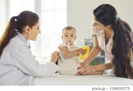 Pediatrician examining infant with mother in clinic, baby holding toy during visit 136630575