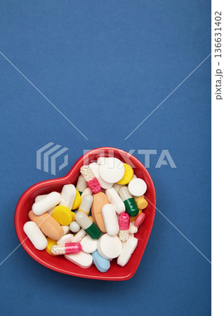 Heart-shaped bowl with a colorful assortment of various supplements on blue background. Health, wellness, and care pharmacology concept. Heart-shaped bowl with a colorful assortment of various supplements on blue background. Health, wellness, and care pharmacology concept. 136631402