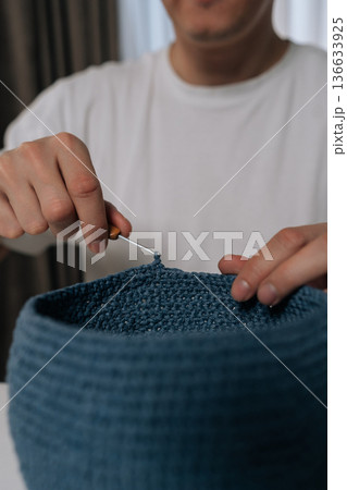 Vertical cropped shot of man enjoying creative hobby, crocheting blue yarn basket at home, hands crafting handmade item with needle and thread, engaging in relaxing and skillful activity, close-up. 136633925