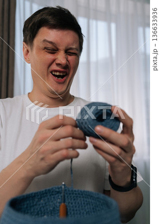 Vertical portrait of funny young man laughing and squinting with joyous emotion while engaged in relaxing hobby of crocheting, holding ball of blue yarn working on textile project with crochet hook. Vertical portrait of funny young man laughing and squinting with joyous emotion while engaged in relaxing hobby of crocheting, holding ball of blue yarn working on textile project with crochet hook. 136633936