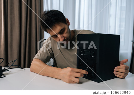Focused young man inserting power cord into back of black studio monitor speaker, ensuring secure connection for audio setup on clean white table. Focused young man inserting power cord into back of black studio monitor speaker, ensuring secure connection for audio setup on clean white table. 136633940
