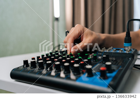 Hand adjusting various knobs on sound mixer console, controlling audio levels and effects for sound engineering, music production, podcasting, or broadcasting in professional home studio setup. Hand adjusting various knobs on sound mixer console, controlling audio levels and effects for sound engineering, music production, podcasting, or broadcasting in professional home studio setup. 136633942