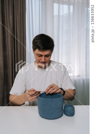 Vertical portrait of young man focused on crocheting blue yarn basket at home, practicing traditional needlework and enjoying mindful, creative hobby that blends skill, relaxation, handmade design. Vertical portrait of young man focused on crocheting blue yarn basket at home, practicing traditional needlework and enjoying mindful, creative hobby that blends skill, relaxation, handmade design. 136633952