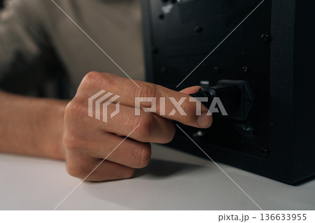 Close-up hand of unrecognizable sound engineer male connecting power cable to black monitor speaker, setting up audio equipment on white desk, preparing home studio or entertainment system for use. 136633955