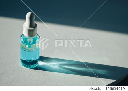 Skincare bottle with blue liquid standing on white surface casting shadow 136634019