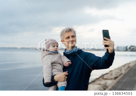 Smiling father holding baby and taking selfie together during outdoors walk. They are enjoying the moment. Parenthood and fatherhood concept. Everyday parenting lifestyle. 136634501