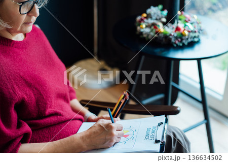 Woman drawing work-life balance wheel sitting on armchair at cozy dark home interior. Self-reflection, life planning for next new year. Personal Year review. Coaching tools. Finding Balance in Life Woman drawing work-life balance wheel sitting on armchair at cozy dark home interior. Self-reflection, life planning for next new year. Personal Year review. Coaching tools. Finding Balance in Life 136634502