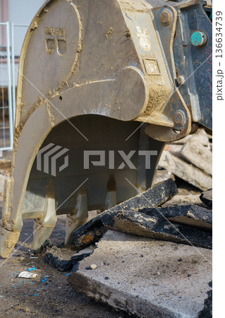 Close-up of hydraulic excavator bucket tearing asphalt and concrete during demolition works. Heavy machinery at urban construction site. Close-up of hydraulic excavator bucket tearing asphalt and concrete during demolition works. Heavy machinery at urban construction site. 136634739