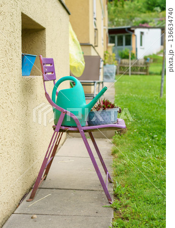 Green watering can and potted succulents on a rustic chair near house wall. Quiet backyard scene with gardening tools and fresh lawn. Green watering can and potted succulents on a rustic chair near house wall. Quiet backyard scene with gardening tools and fresh lawn. 136634740