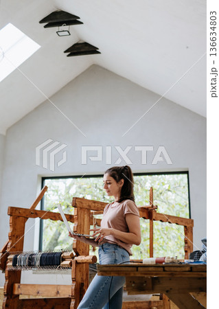 Young businesswoman checking customer order on laptop, standing by wooden loom. Upcycled textile, small weaving business and circular economy. Young businesswoman checking customer order on laptop, standing by wooden loom. Upcycled textile, small weaving business and circular economy. 136634803