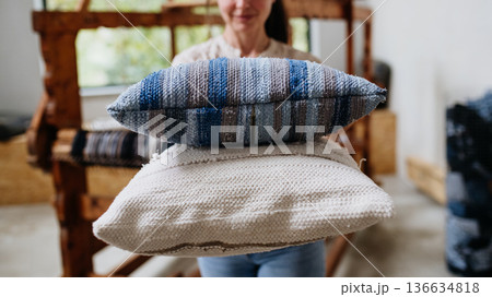 Young businesswoman next to wooden loom, holding pillows with woven covers made from recycled fabrics. Upcycling in business and circular economy. Young businesswoman next to wooden loom, holding pillows with woven covers made from recycled fabrics. Upcycling in business and circular economy. 136634818