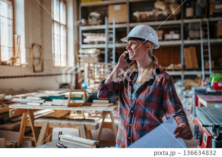 Female woodworker making phone call with client, standing in carpentry workshop. 136634832