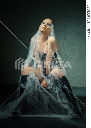 Dramatic portrait of a bald woman in a black corset and boots trapped in spiderwebs. 136635989