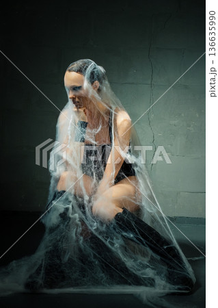 Conceptual portrait of a woman ensnared in a cocoon of spiderwebs against a dark wall. Conceptual portrait of a woman ensnared in a cocoon of spiderwebs against a dark wall. 136635990