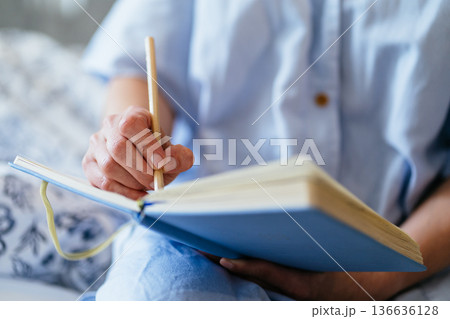 close up, middle aged White woman hand holding pencil, writing in blue notebook on bed, soft natural light, cozy home bedroom, mindful journaling, personal goals, quiet self care moment. 136636128