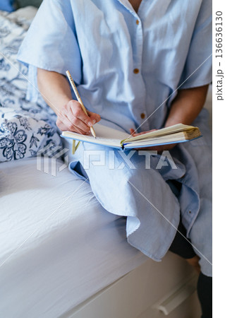 Vertical close up, unrecognizable middle aged White woman hands holding pencil, writing in open blue notebook on bed, linen shirt dress, cozy bedroom textile, personal planning, mindfulness practice Vertical close up, unrecognizable middle aged White woman hands holding pencil, writing in open blue notebook on bed, linen shirt dress, cozy bedroom textile, personal planning, mindfulness practice 136636130