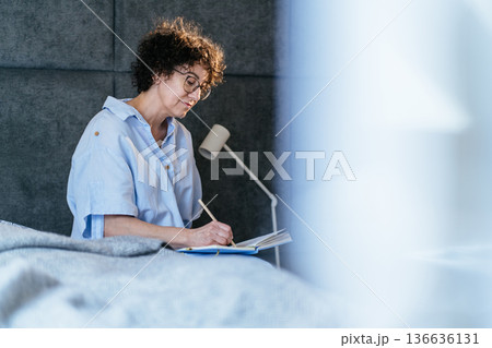 intelligent middle aged woman, curly hair, glasses, journaling on bed, writing in blue notebook, pencil, bedroom lamp, mindful morning routine, self care, cozy home lifestyle, ritual, morning pages intelligent middle aged woman, curly hair, glasses, journaling on bed, writing in blue notebook, pencil, bedroom lamp, mindful morning routine, self care, cozy home lifestyle, ritual, morning pages 136636131