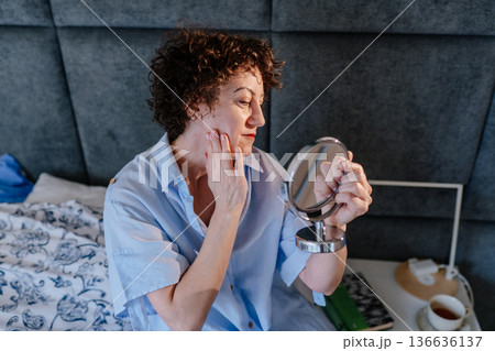 Curly-haired mature woman in blue pajamas looking at her reflection with a satisfied smile. Concept of radiant skin, self-appreciation, and pensive yet happy morning rituals at home. 136636137