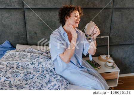 well-groomed curly-haired mature woman in blue pajamas sits on a bed, applying moisturizer to her neck. Concept of anti-aging skincare, pensive morning rituals, and authentic domestic beauty. well-groomed curly-haired mature woman in blue pajamas sits on a bed, applying moisturizer to her neck. Concept of anti-aging skincare, pensive morning rituals, and authentic domestic beauty. 136636138