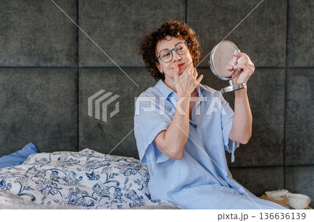 Serene and thoughtful woman in blue pajamas looking in a hand mirror with a gentle smile while touching her face. Concept of self-appreciation, effective skincare, and radiant aging in a cozy home. Serene and thoughtful woman in blue pajamas looking in a hand mirror with a gentle smile while touching her face. Concept of self-appreciation, effective skincare, and radiant aging in a cozy home. 136636139