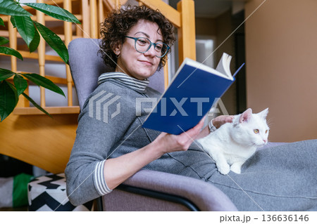 A curly-haired woman in glasses relaxes in a comfortable armchair at home, reading a book and petting her white cat. Concept of domestic peace, animal companionship, and slow living lifestyle. 136636146