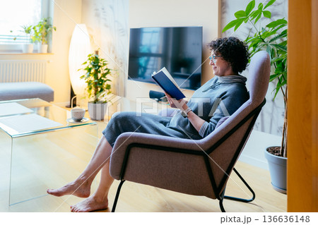 High angle of a mature woman in a blue armchair reading a book. Cozy home interior, hot tea, slow living and mindfulness concept. Mental health, education and peaceful weekend in modern apartment. 136636148