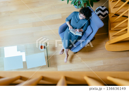 High angle shot of a woman relaxing in a stylish blue armchair, reading a book with a cup of tea nearby. Concept of slow living, education, hobby, and peaceful weekend at home. High angle shot of a woman relaxing in a stylish blue armchair, reading a book with a cup of tea nearby. Concept of slow living, education, hobby, and peaceful weekend at home. 136636149