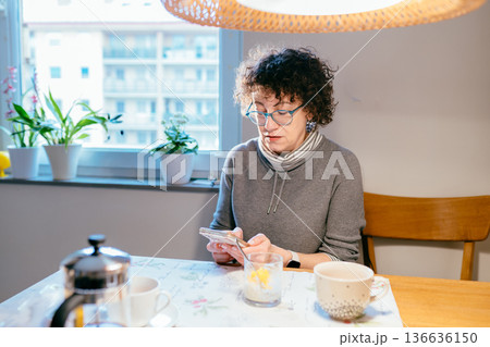 Attractive mature woman using a mobile phone while sitting at a kitchen table with tea and breakfast. Modern technology, social media addiction, or morning news check-up concept. Attractive mature woman using a mobile phone while sitting at a kitchen table with tea and breakfast. Modern technology, social media addiction, or morning news check-up concept. 136636150