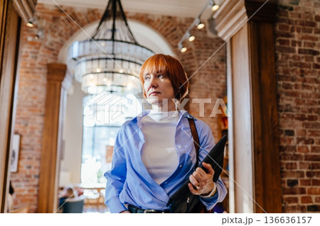 Confident businesswoman walking inside modern cafe holding document folder. Thoughtful expression Urban public space, brick interior, daylight, leadership concept, modern professional lifestyle Confident businesswoman walking inside modern cafe holding document folder. Thoughtful expression Urban public space, brick interior, daylight, leadership concept, modern professional lifestyle 136636157