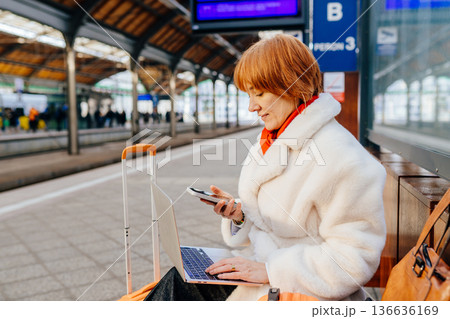 Experienced caucasian redhead professional using smartphone and laptop at train station platform. Managing communication and tasks during travel, hybrid work routine, in public transport environment. Experienced caucasian redhead professional using smartphone and laptop at train station platform. Managing communication and tasks during travel, hybrid work routine, in public transport environment. 136636169