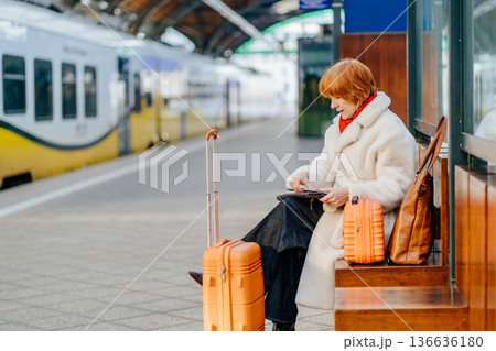 Caucasian redhead adult woman sitting on wooden bench at railway station, using smartphone, waiting for train, orange suitcase, white coat, red scarf, winter indoor platform , solo travel routine Caucasian redhead adult woman sitting on wooden bench at railway station, using smartphone, waiting for train, orange suitcase, white coat, red scarf, winter indoor platform , solo travel routine 136636180