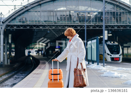 Caucasian redhead adult woman standing on railway platform holding orange suitcase, white coat and red scarf, modern electric train in background, winter urban rail travel, journey pause departure Caucasian redhead adult woman standing on railway platform holding orange suitcase, white coat and red scarf, modern electric train in background, winter urban rail travel, journey pause departure 136636181