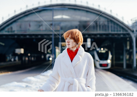 Caucasian redhead adult woman standing on railway platform, white coat and red scarf, thoughtful attentive expression, modern train in background, winter city rail travel pause 136636182
