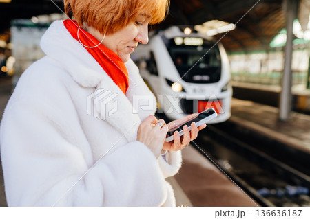 Close view of caucasian redhead adult woman holding smartphone on railway station platform, white coat and red scarf, checking information before train arrival, digital travel routine Close view of caucasian redhead adult woman holding smartphone on railway station platform, white coat and red scarf, checking information before train arrival, digital travel routine 136636187