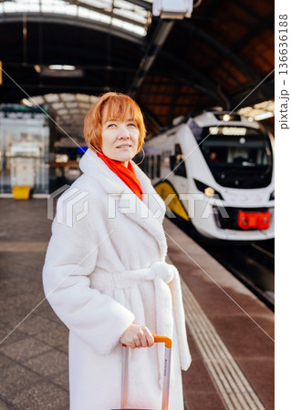 Vertical portrait of mature woman standing on railway platform, holding orange suitcase, white coat and red scarf, modern electric train behind, winter urban rail mobility, solo travel decision moment Vertical portrait of mature woman standing on railway platform, holding orange suitcase, white coat and red scarf, modern electric train behind, winter urban rail mobility, solo travel decision moment 136636188