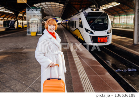Caucasian redhead adult woman standing on railway station platform with orange suitcase, white coat and red scarf, looking at camera, modern electric train behind, travel, winter urban mobility. Caucasian redhead adult woman standing on railway station platform with orange suitcase, white coat and red scarf, looking at camera, modern electric train behind, travel, winter urban mobility. 136636189