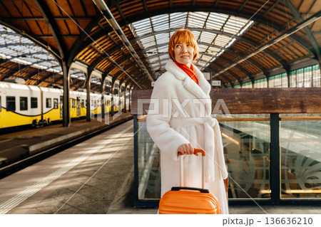 Caucasian redhead adult woman standing on railway station platform beside glass wall, reflection visible, holding orange suitcase, white fur coat and red scarf, eyes closed, sustainable travel concept 136636210