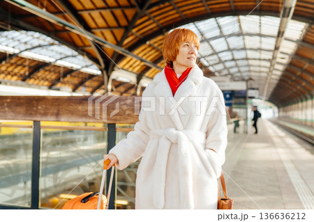 Caucasian redhead adult woman standing on railway station platform, with orange suitcase, white fur coat, red scarf, looking aside, winter travel, mobility, eco transport, journey, departure moment Caucasian redhead adult woman standing on railway station platform, with orange suitcase, white fur coat, red scarf, looking aside, winter travel, mobility, eco transport, journey, departure moment 136636212