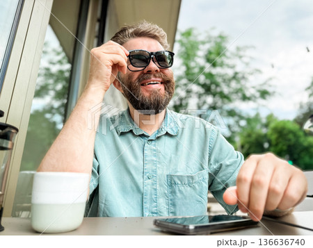 Man smiling while wearing clip on sunglasses at outdoor cafe table. Glasses with sunglasses magnetic clip. Man smiling while wearing clip on sunglasses at outdoor cafe table. Glasses with sunglasses magnetic clip. 136636740