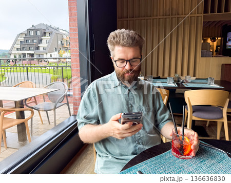 Man sitting at restaurant table holding smartphone and cocktail. Leisure, lifestyle, and hospitality atmosphere with social interaction and guest experience. Man sitting at restaurant table holding smartphone and cocktail. Leisure, lifestyle, and hospitality atmosphere with social interaction and guest experience. 136636830