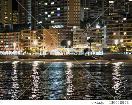 City Lights Reflect On Tranquil Waters, Lights From City Buildings Dance On Serene Ocean Surface At Night 136638998