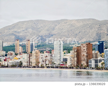 Coastal Cityscape With Tranquil Waters, Calm Shoreline Towers Reflected Beneath Clouded Morning Sky 136639020