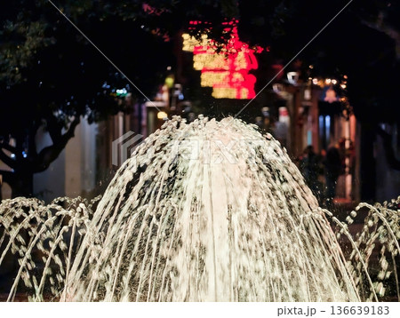 Fountain Under Night, Central Nozzle Creates Illuminated Symmetrical Water Display In Urban Setting Fountain Under Night, Central Nozzle Creates Illuminated Symmetrical Water Display In Urban Setting 136639183