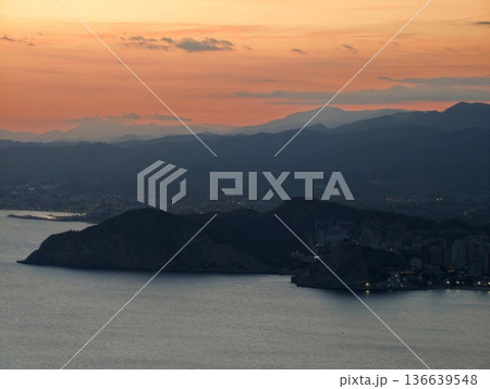 Dramatic Silhouette Of Headland Against Sunset Sky With Peaceful Bay And Tiny City Lights Along Shoreline 136639548